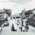 Black and white photo of Women and various ages of children wearing white aprons and hats standing on the High Street of Drayton St Leonard